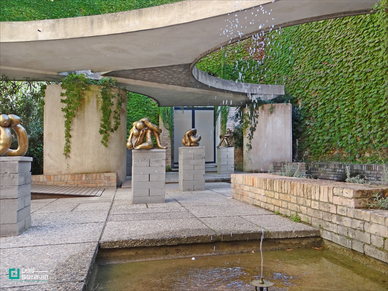 نمایی از «Giardino delle Sculture» کارلو اسکارپا در بینال ونیز؛ فضای حیاطی با سقف بتنی منحنی، دیوارهای پوشیده از گیاهان سبز، حوضچه آب و آب‌نما در پیش‌زمینه و مجسمه‌های برنزی روی پایه‌های بتنی در میان محوطه.