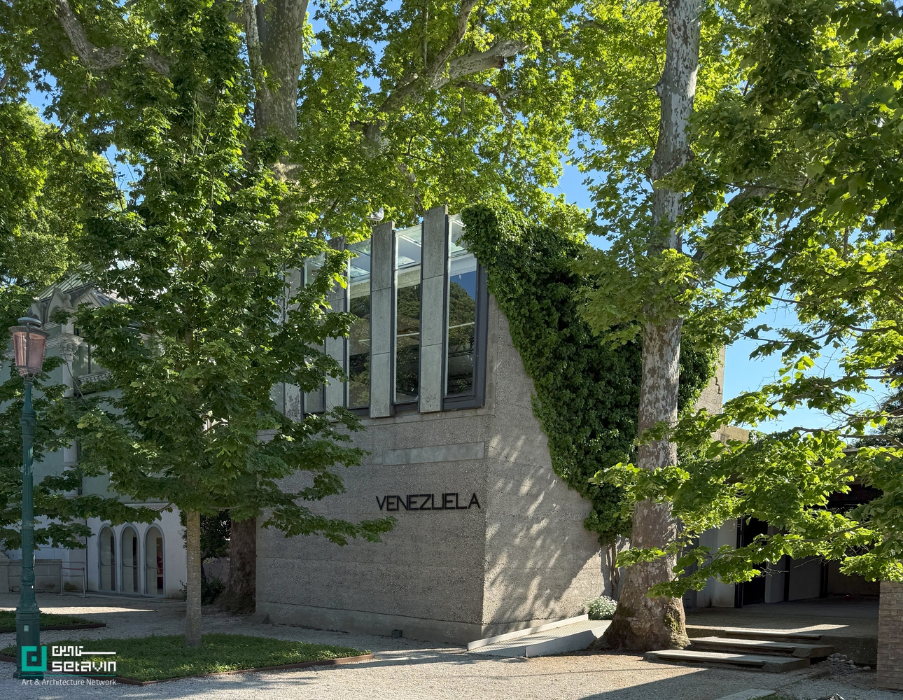 نمای بیرونی پاویون ونزوئلا در باغ‌های جاردینی بینال ونیز، اثر کارلو اسکارپا؛ حجم بتنی با پنجره‌های عمودی شیشه‌ای، پوشیده از گیاهان بالارونده در میان درختان بلند، با نام VENEZUELA روی نما.
