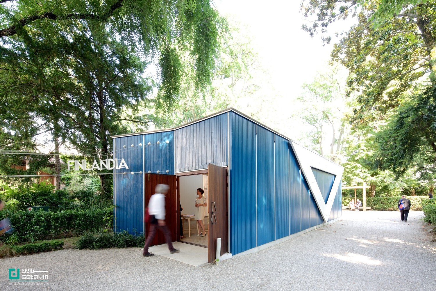 ۲. نمای بیرونی پاویون فنلاند در باغ‌های جاردینی بینال ونیز، اثر آلوار آلتو و الیسّا آلتو؛ حجم موقت با بدنه فلزی آبی‌رنگ، ورودی چوبی باز، تابلو روشن «FINLANDIA» و بازدیدکنندگان در حال حرکت در میان فضای سبز.
