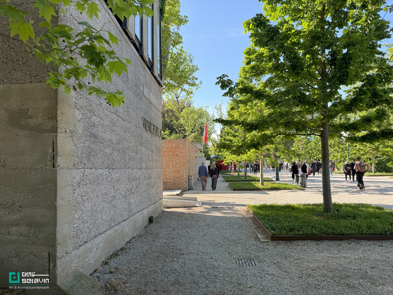 نمای جانبی پاویون ونزوئلا در باغ‌های جاردینی بینال ونیز؛ دیوار بتنی با نام VENEZUELA در سمت چپ و مسیر پیاده‌روی درختکاری‌شده با حضور بازدیدکنندگان در سمت راست تصویر.