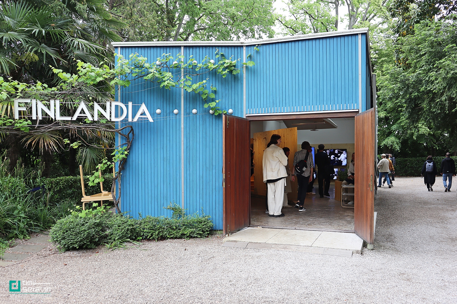 نمای نزدیک ورودی پاویون فنلاند در باغ‌های جاردینی بینال ونیز؛ جداره فلزی آبی با لوگوی «FINLANDIA»، درهای چوبی باز و گروهی از بازدیدکنندگان که در حال ورود و تماشای نمایشگاه داخل پاویون هستند، در میان پوشش گیاهی سبز اطراف.
